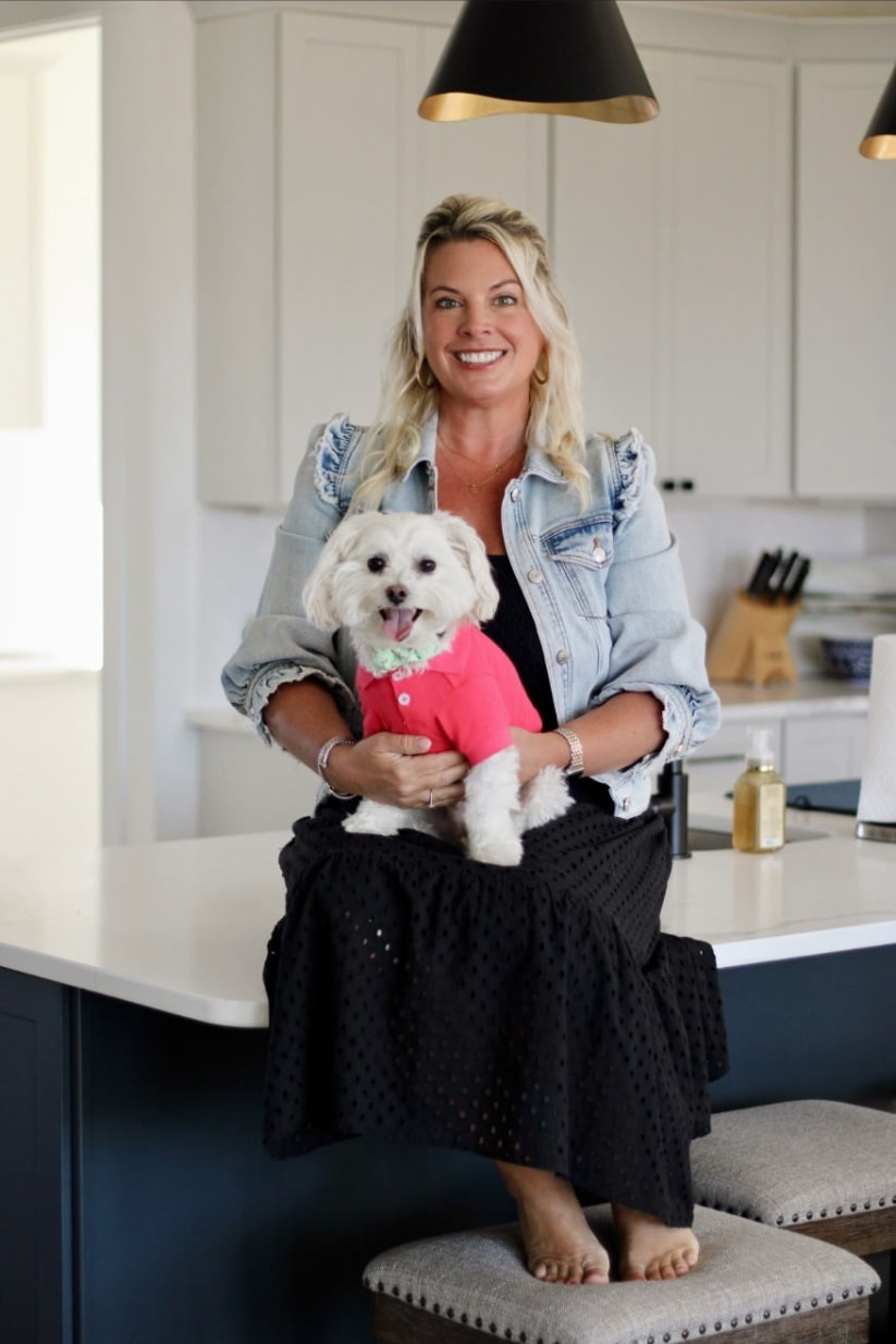 Casey Styers smiling in the kitchen while holding Rocky, her white dog in a pink shirt